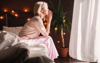 A contemplative woman in a pink nightgown sits on the bed, hands together in reflection, gazing toward a softly-lit window with a potted plant in the background, creating a tranquil atmosphere. She is managing emotional stress.