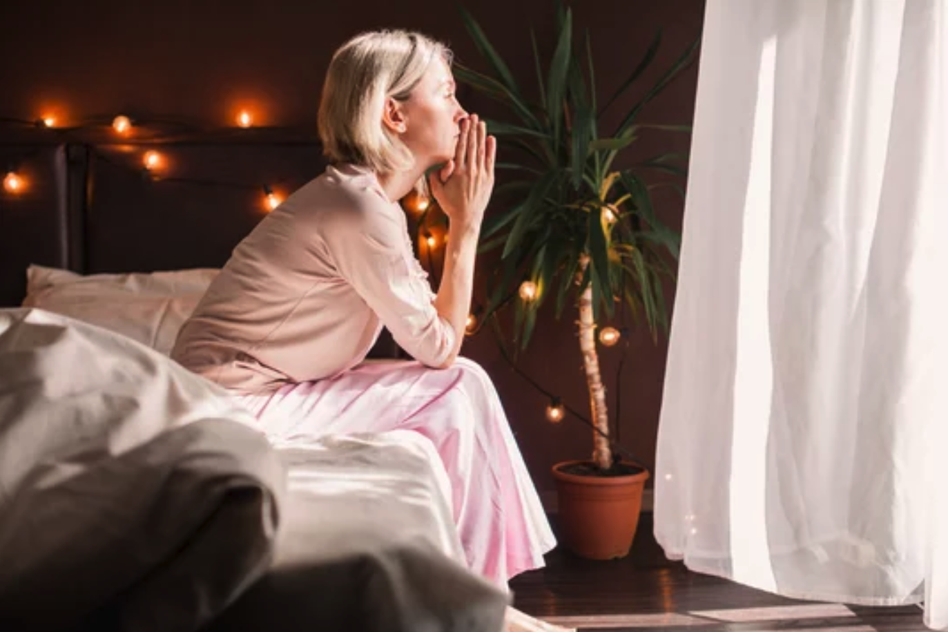 A contemplative woman in a pink nightgown sits on the bed, hands together in reflection, gazing toward a softly-lit window with a potted plant in the background, creating a tranquil atmosphere. She is managing emotional stress.