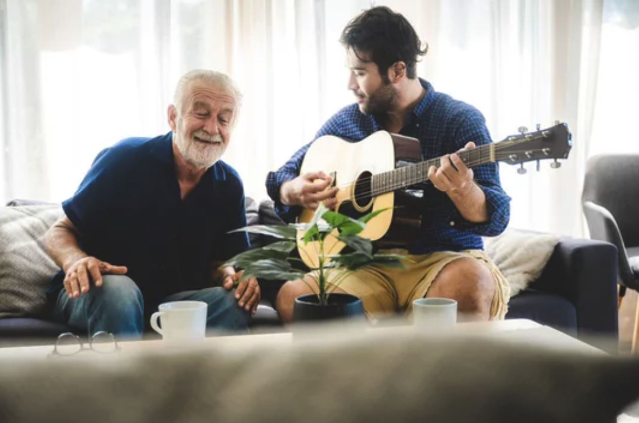 A young man playing guitar for an elderly man who is smiling and clapping along.