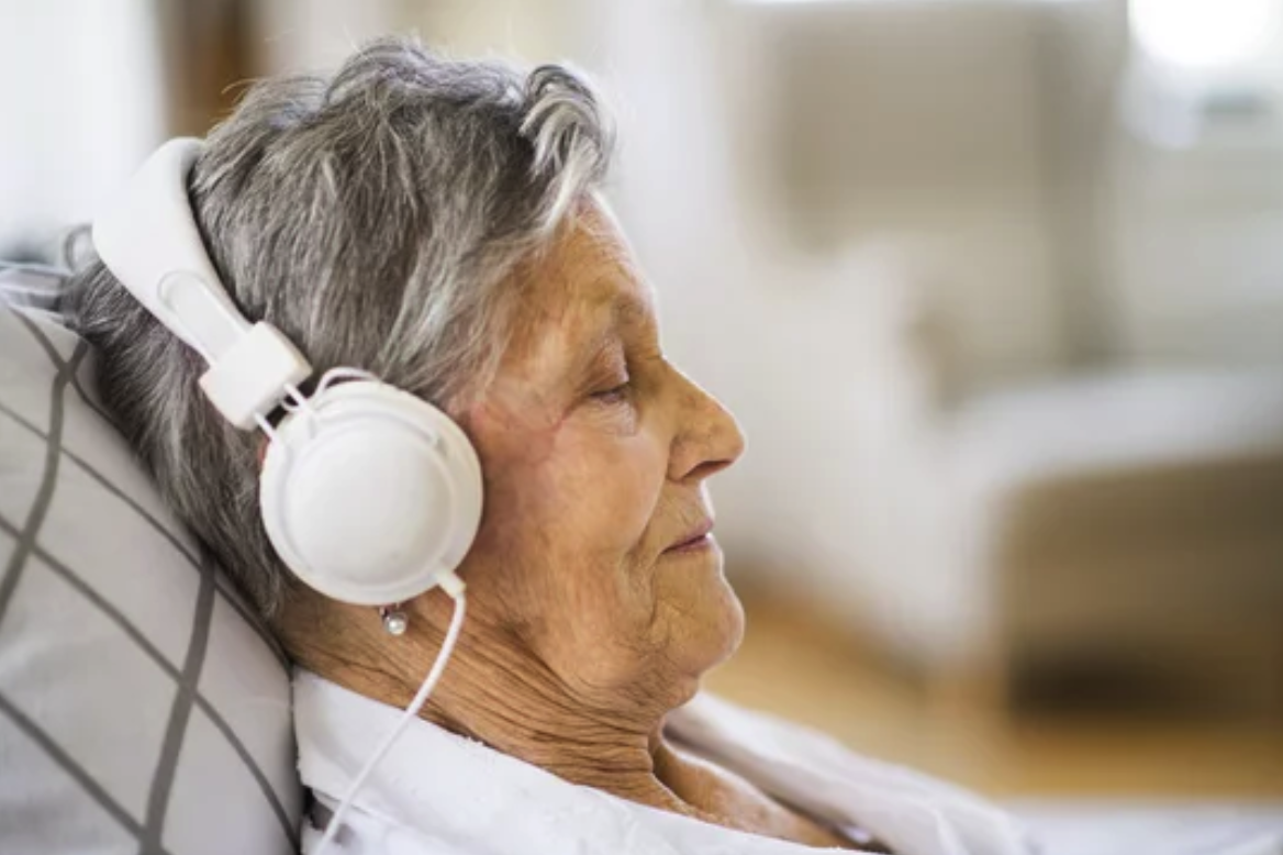 An elderly woman resting in a chair, enjoying music through white headphones.