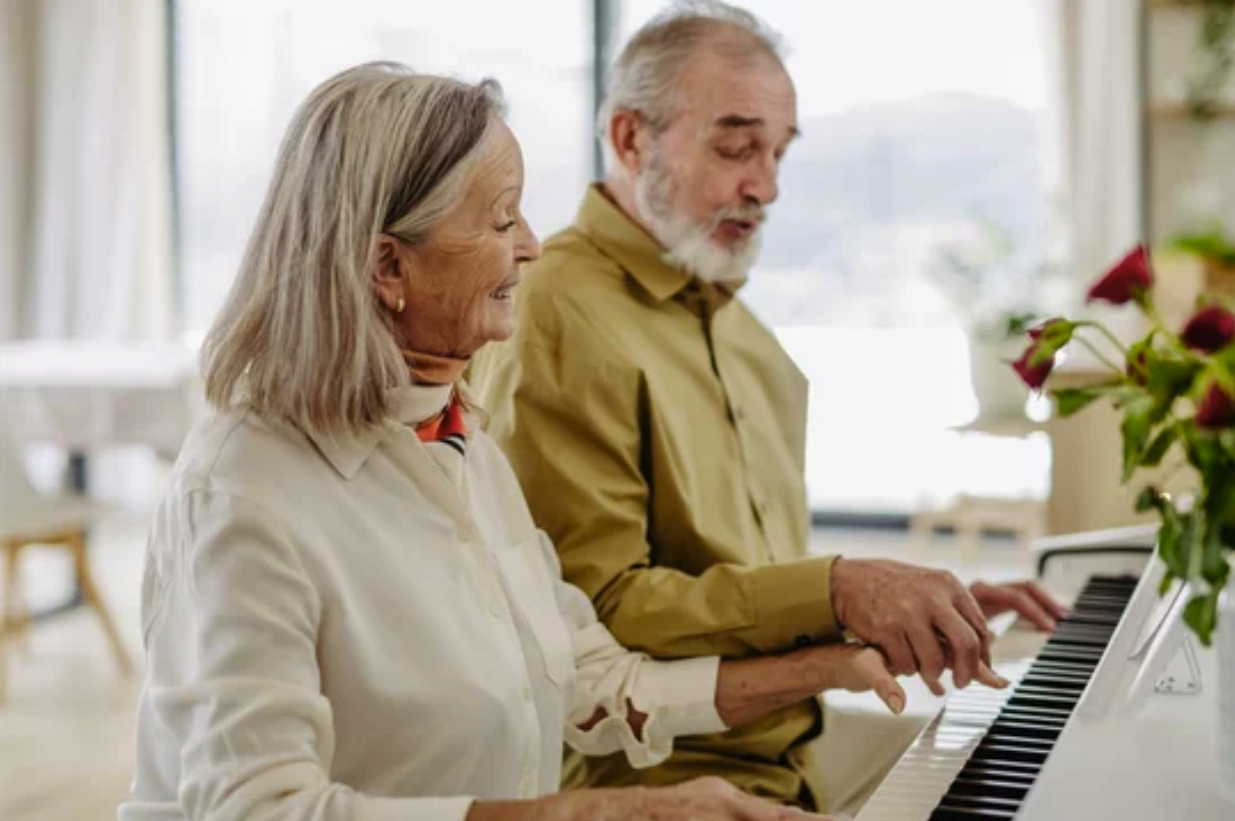 An elderly couple playing the piano together in a bright room.