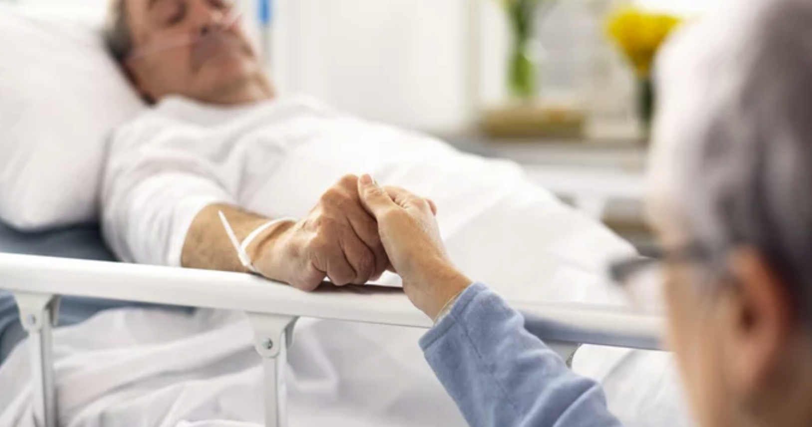 Two people holding hands, one lying in a hospital bed and the other sitting beside them, offering comfort.